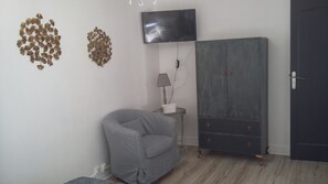 Um quarto com uma poltrona cinza, um armário com gavetas, uma TV de tela plana presa na parede e um abajur sobre uma mesa pequena.