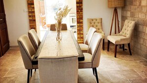 A dining area with a wooden table, six chairs, and a vase with dried plants. An arched brick wall and a lamp are also visible.