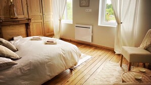 A bedroom with a wooden bed, white linens, a chair, a small table, and a window with curtains.