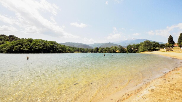 Un lac aux eaux limpides, bordé d’une plage de sable, entouré d’une végétation luxuriante et avec des montagnes en toile de fond.