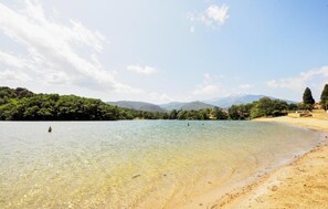 Ein klarer See mit Sandstrand, umgeben von üppiger Vegetation und Bergen im Hintergrund.