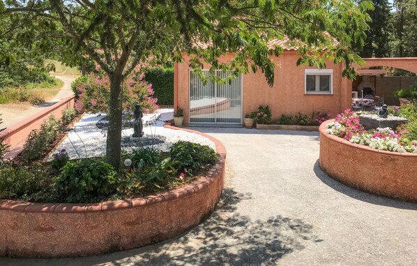 Un chemin de jardin mène à un bâtiment doté d’une entrée fermée par un portail, le tout étant entouré de parterres de fleurs aménagés et d’un petit espace patio.
