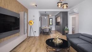 A modern living room with a flat-screen TV, a round coffee table, a grey sofa, and a dining area with a table set for four. - Stunning apartment in Radmani (Radmani)