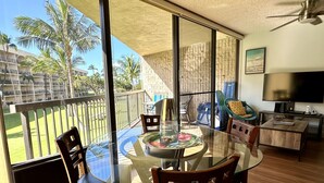 A modern living room with a glass dining table, wooden chairs, a TV, and a balcony with a view of palm trees and a building. - Spacious Maui Sunset Condo: Steps to BEACH. Pool, Gym, AC & Family Friendly (Kihei)