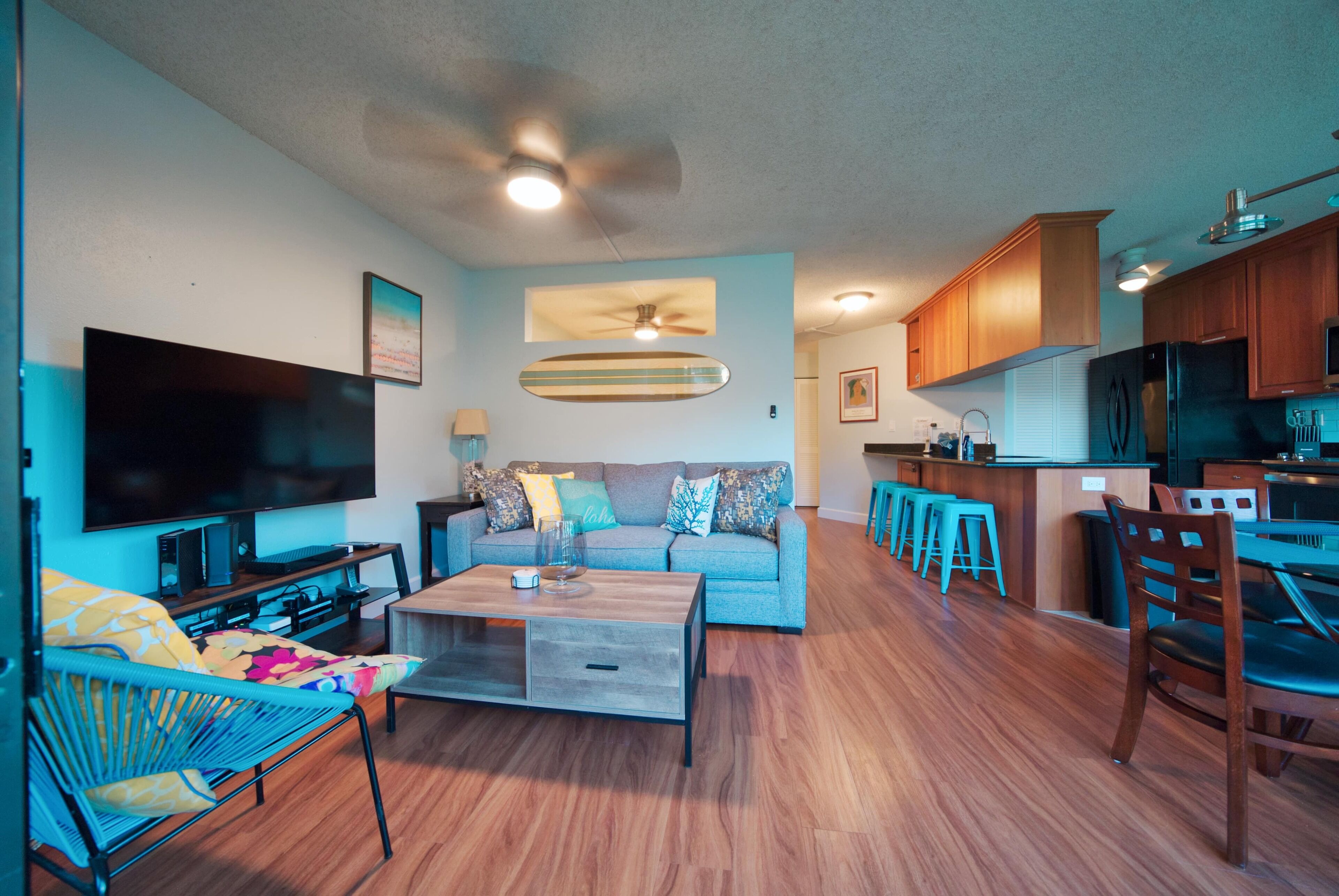 A modern living room with a flat-screen TV, a wooden coffee table, a sofa with colorful cushions, and a kitchen area with wooden cabinets and a refrigerator.