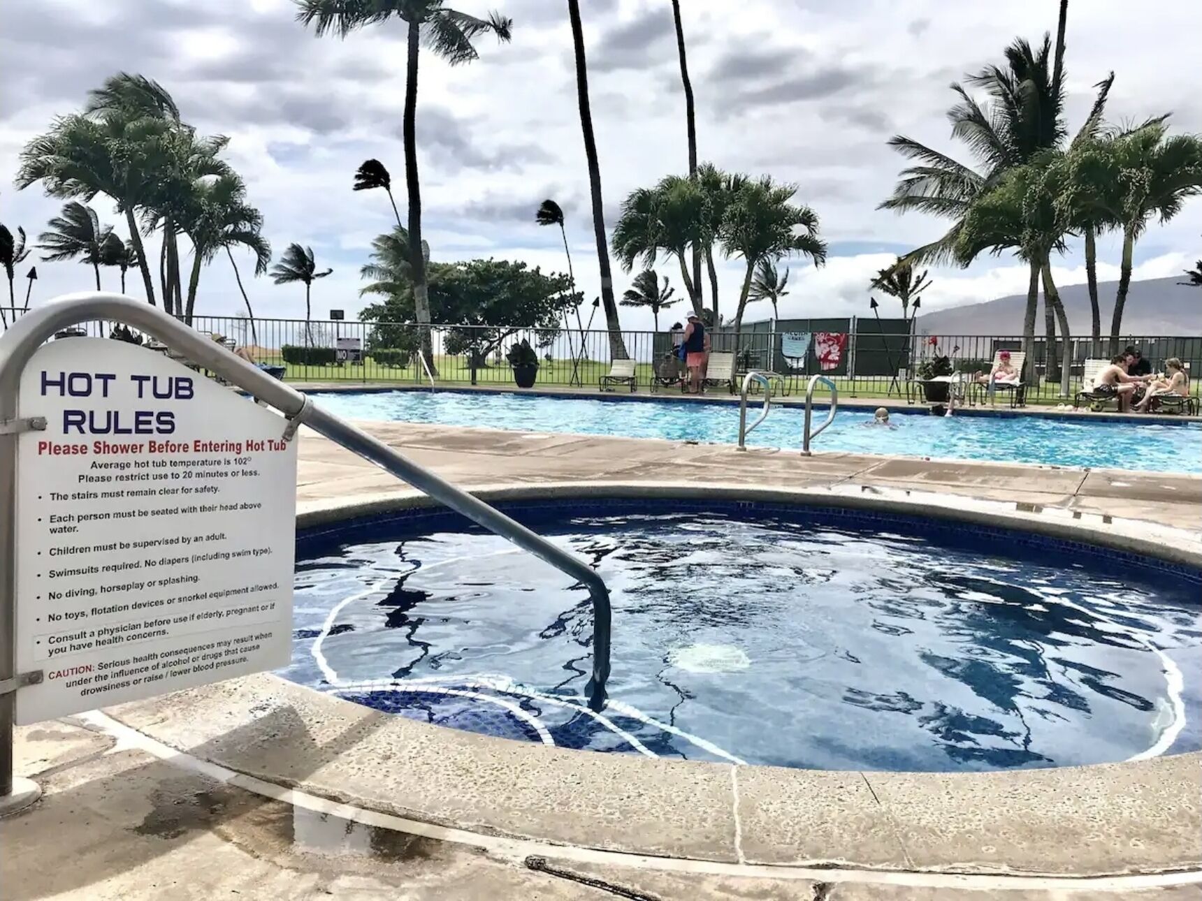 Een zone met een jacuzzi, een bord met regels en een zwembad met palmbomen en mensen.