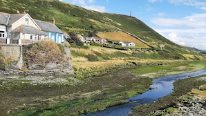 Ein Küstendorf mit Häusern entlang eines Flussufers, einer Steinmauer und einer Hügelkulisse im Hintergrund.