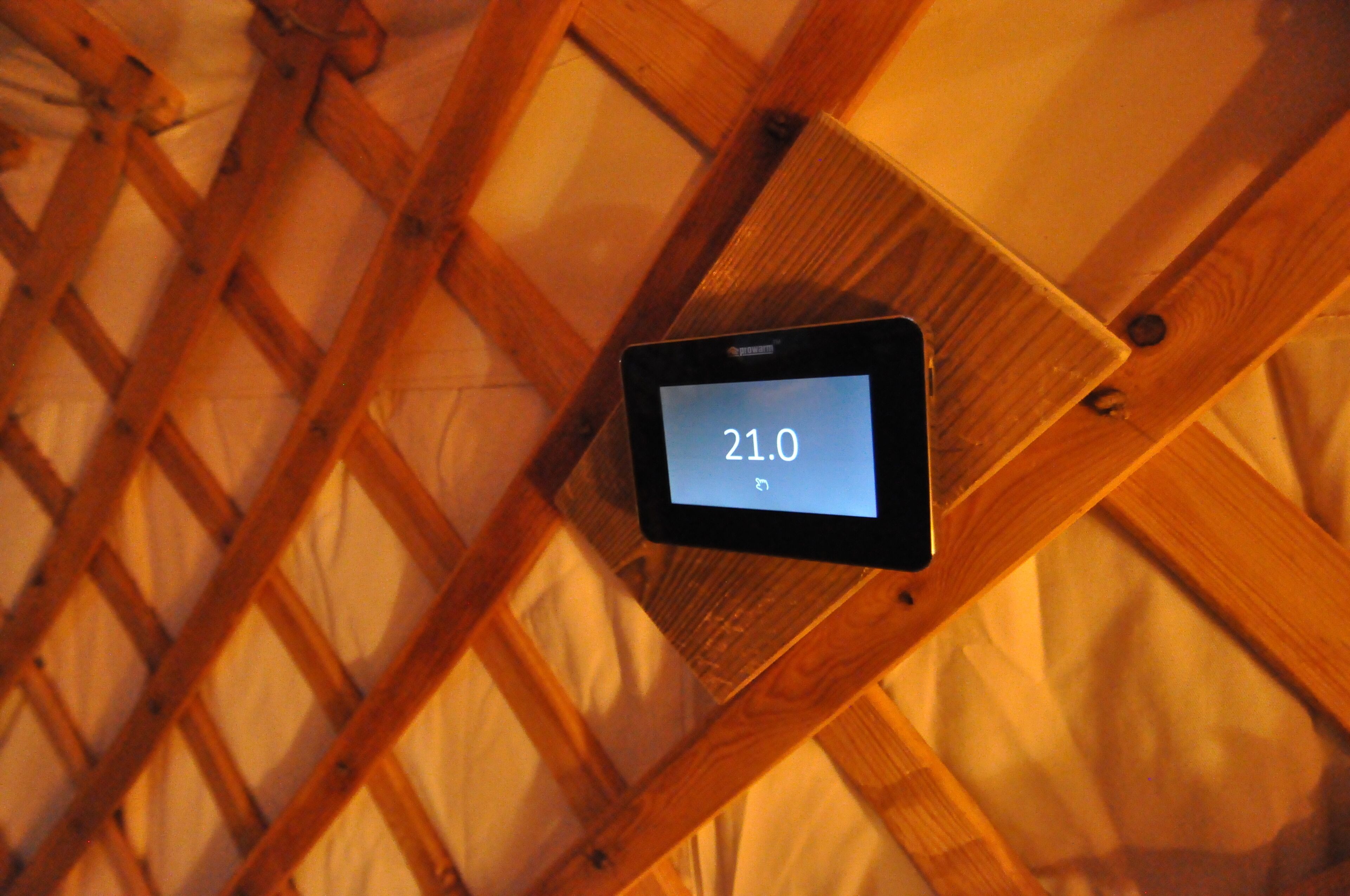 Een tablet toont een temperatuur van 21,0 graden Celsius, bevestigd aan een houten balk in een kamer met een spijlen plafond.