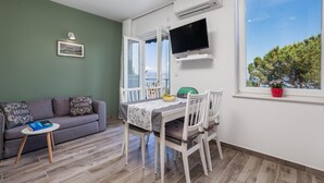 Ein Wohnzimmer mit einer Couch, einem Esstisch mit Stühlen, einem an der Wand befestigten Fernseher und einem Fenster mit Blick auf Bäume.