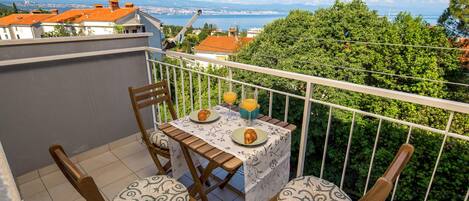 A balcony with a table set for two, two chairs, and a scenic view of a city and sea.