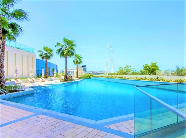 A swimming pool with lounge chairs and palm trees, a Ferris wheel in the background, and modern buildings.