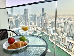 A table with a croissant, an apple, a green apple, and a glass of orange juice, with a cityscape view through a glass window.