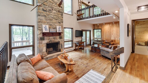 Un vaste espace de vie doté d’une cheminée en pierre, de planchers en bois, d’une mezzanine accessible par un escalier en bois, et de grandes fenêtres offrant une vue sur l’extérieur.