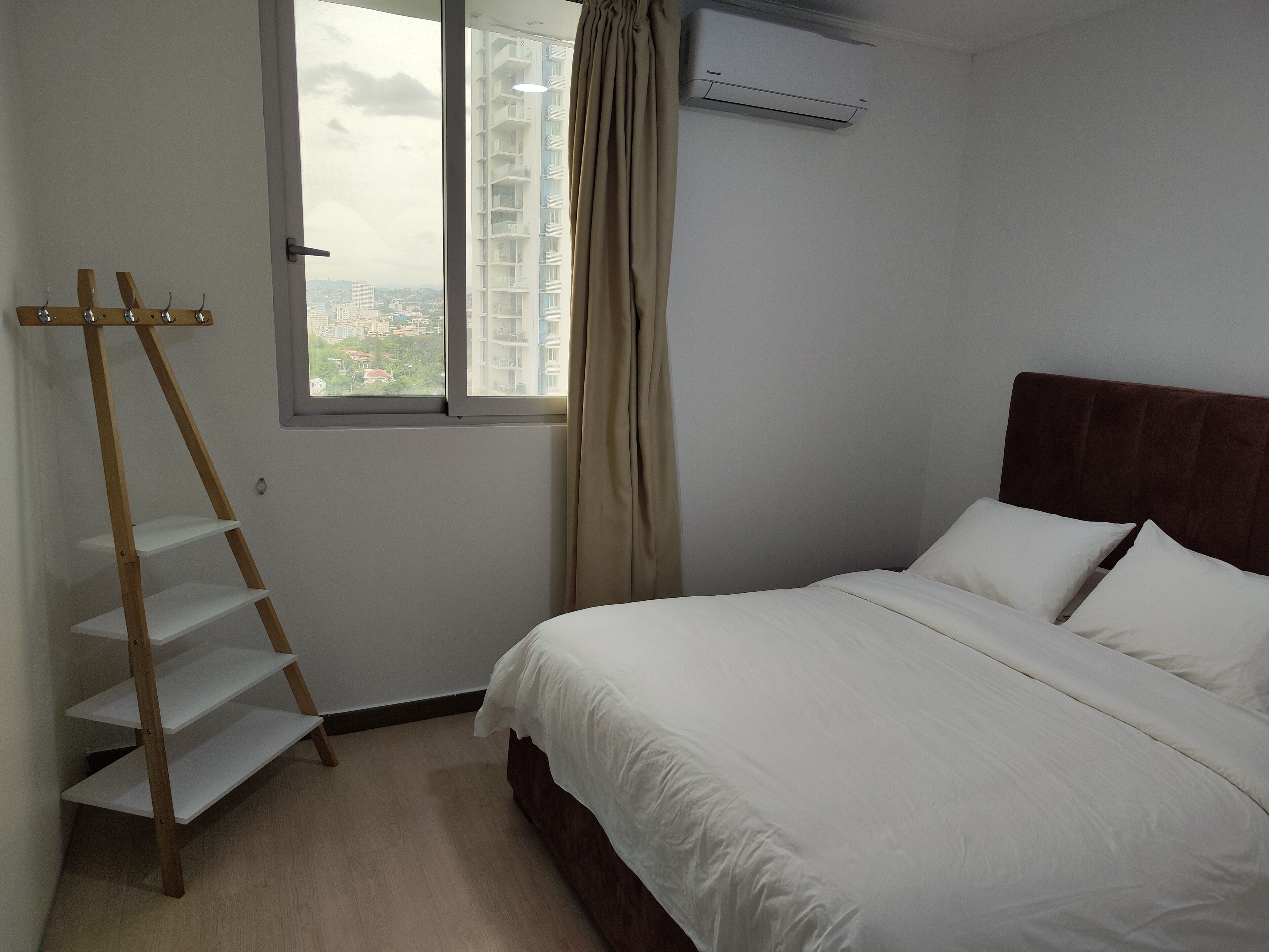 Ein Schlafzimmer mit einem Bett, einem Fenster mit Blick auf Gebäude, einer Holzleiter und einer Klimaanlage.
