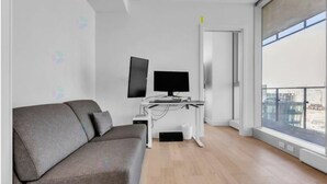 Un salon moderne avec un canapé gris, un téléviseur à écran plat posé sur un meuble blanc et une grande fenêtre donnant sur la ville.