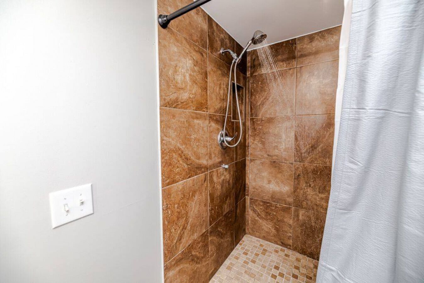 A shower with a handheld showerhead and a wall-mounted faucet, tiled walls, and a white curtain.