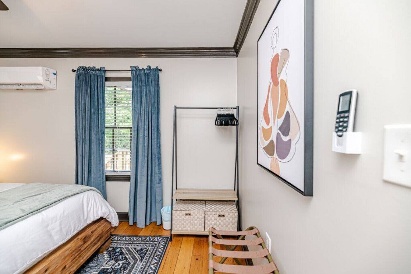 Un dormitorio con cama, una ventana con cortinas, teléfono fijo en la pared, radiador y un cuadro abstracto enmarco.