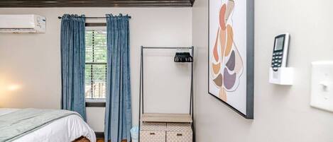 Un dormitorio con cama, una ventana con cortinas, teléfono fijo en la pared, radiador y un cuadro abstracto enmarco.