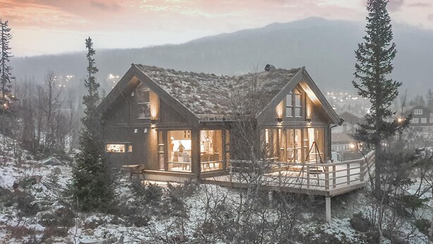 A two-story wooden cabin with a balcony, surrounded by snow-covered trees and mountains in the background.