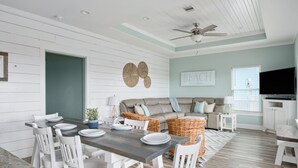 Ein Essbereich mit Holztisch und weißen Stühlen, eine Couch mit Kissen, ein Deckenventilator und ein Schild mit der Aufschrift 'BEACH'.