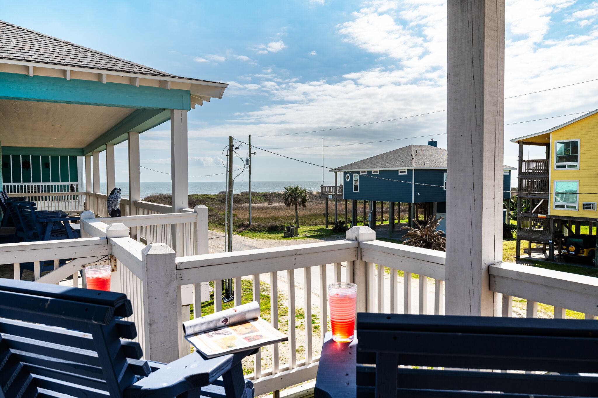 A covered outdoor area with a view of the beach, featuring a wooden deck, chairs, and a table with a drink.