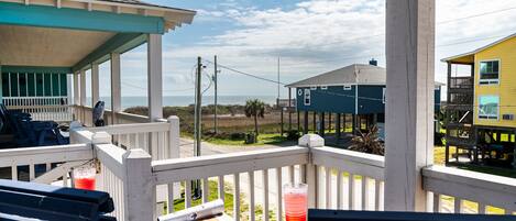 A covered outdoor area with a view of the beach, featuring a wooden deck, chairs, and a table with a drink.