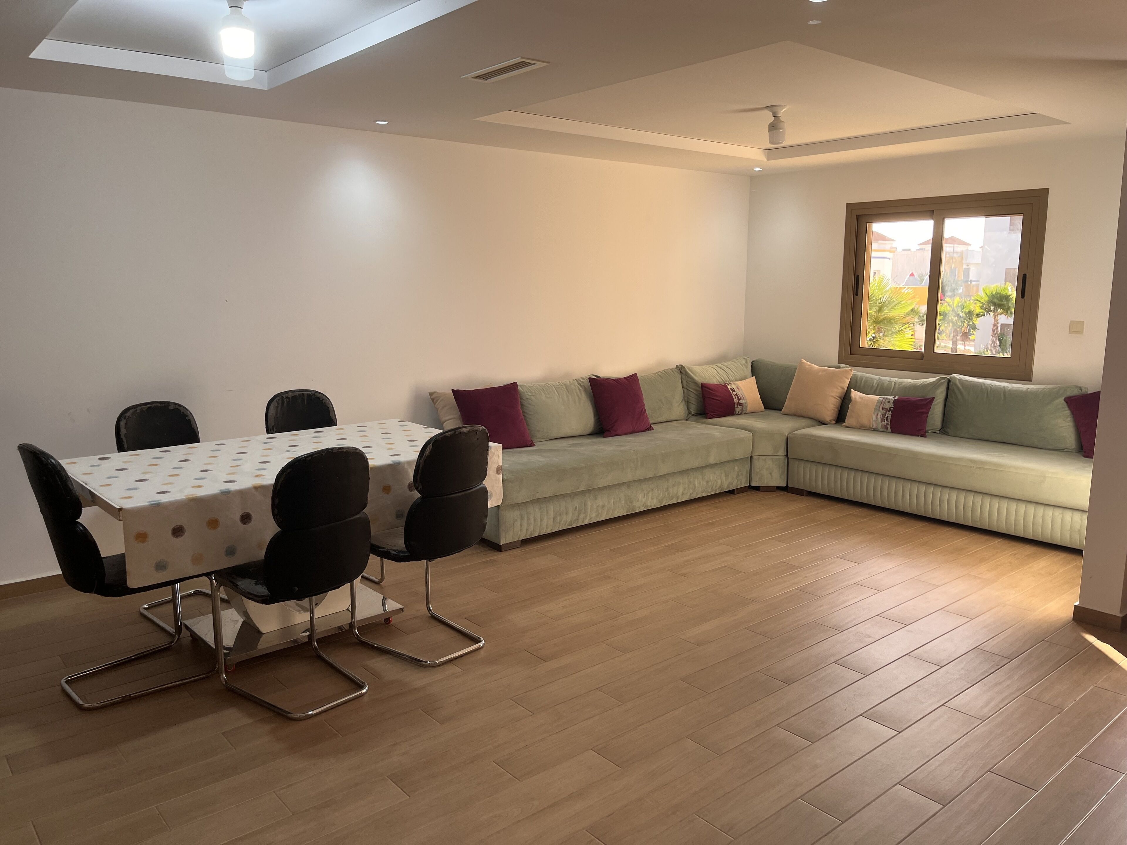 A modern living room with a dining table, black chairs, a green sofa with cushions, and a large window.