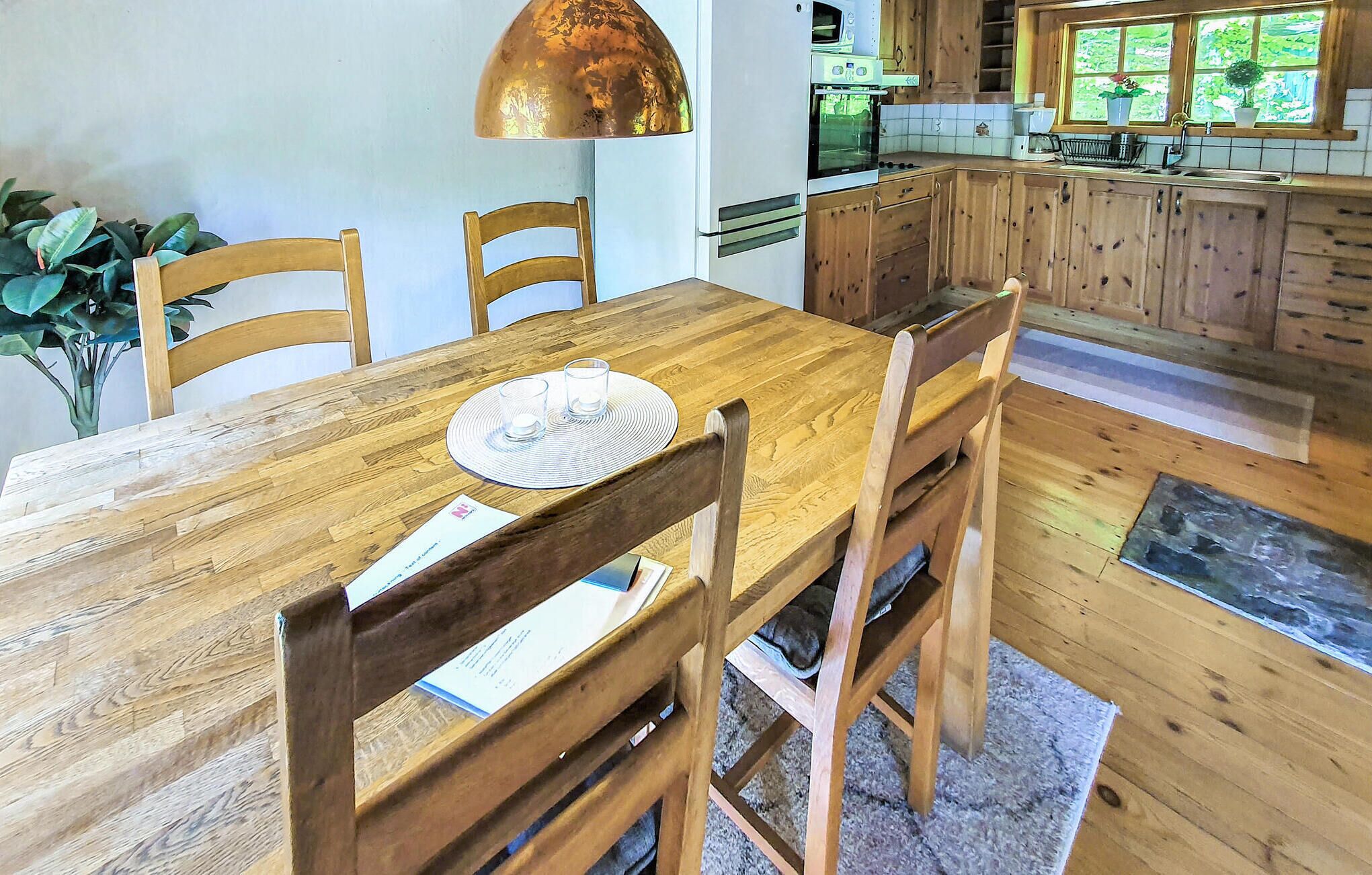 Una mesa de comedor de madera con sillas, una lámpara colgante y una cocina al fondo.