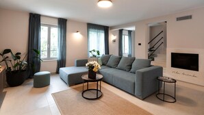 A modern living room with a grey sofa, a small round table with a vase of flowers, a large window with curtains, and a fireplace.