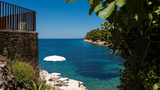 A coastal view with a clear blue sea, a stone wall, a metal railing, and a white umbrella.