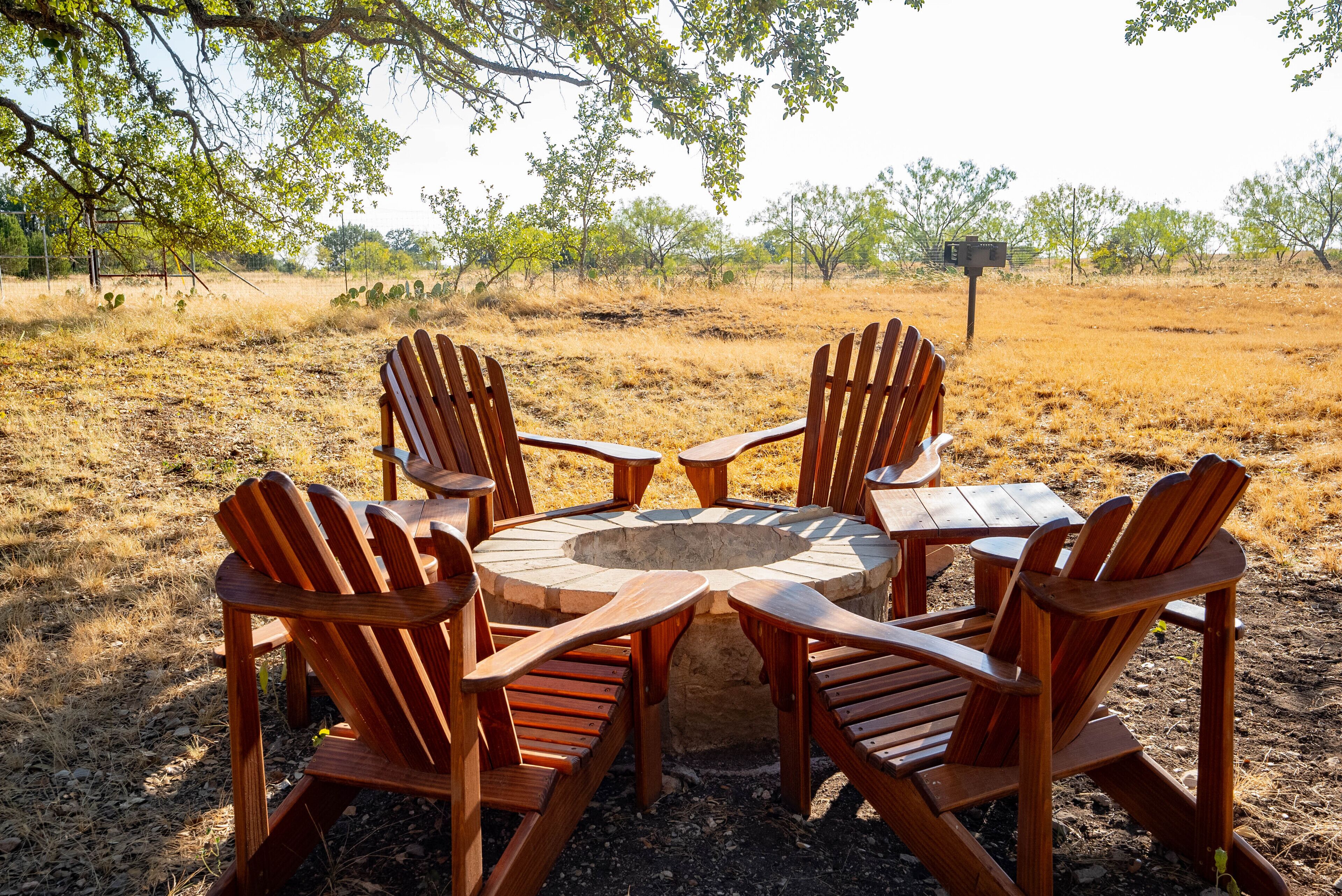 Une table ronde en bois, entourée de quatre chaises en bois, est placée dans un champ ouvert où la végétation est clairsemée et le ciel est dégagé.