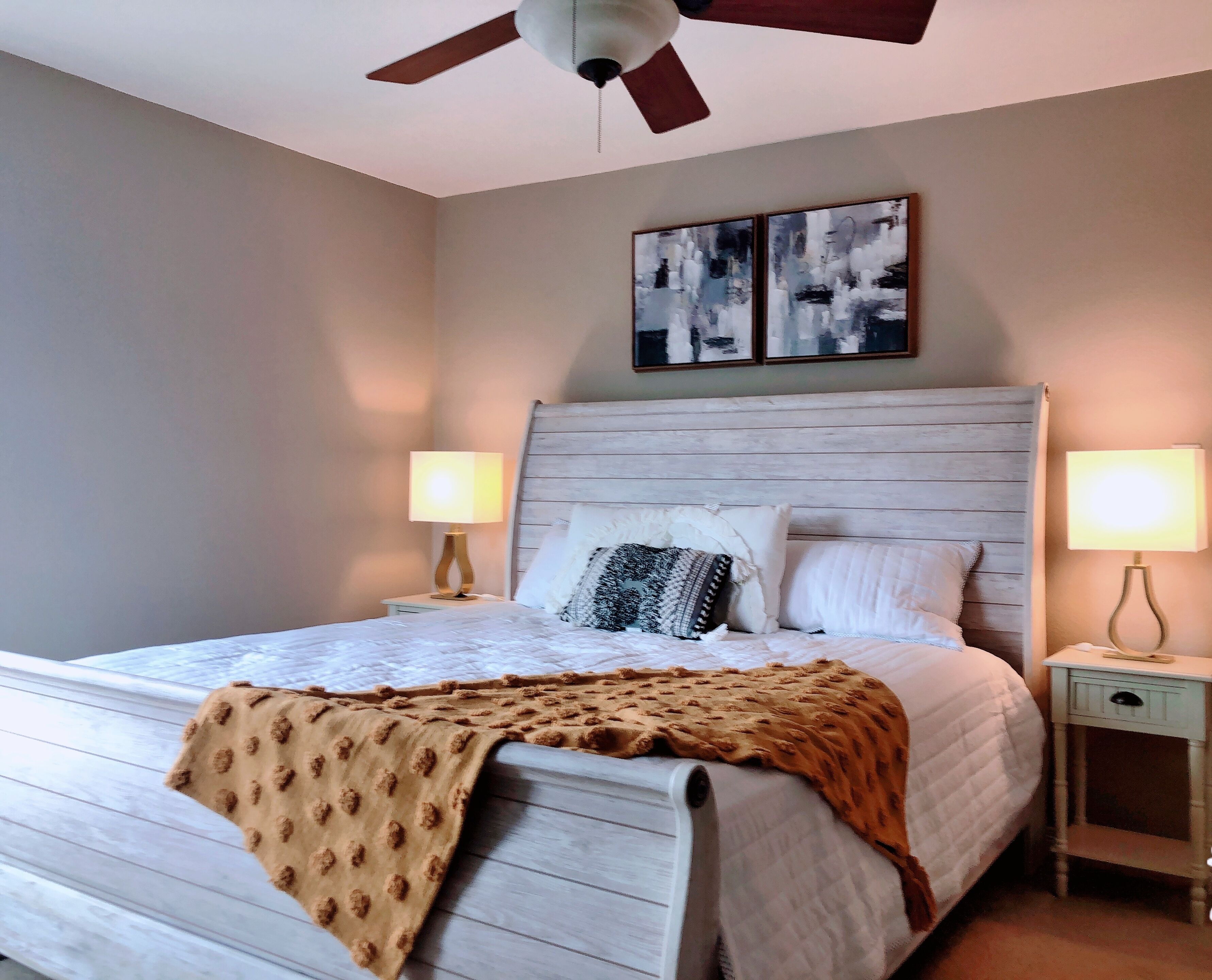 A neatly made bed with a white duvet and a brown throw blanket, flanked by two bedside tables with lamps, a ceiling fan, and abstract wall art above the bed.