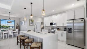 A modern kitchen with white cabinets, stainless steel appliances, a large island with stools, pendant lights, and a view to the outside. - Beautiful waterfront home with heated salt water pool, outdoor kitchen (Cape Coral)