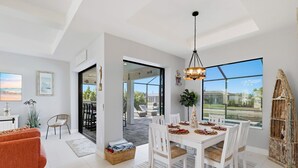 Ampla sala de estar com mesa de jantar, cadeiras brancas e uma grande janela com vista para o exterior.
