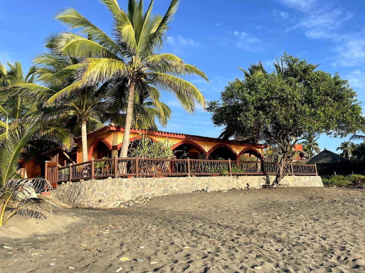 A beachside building with a stone wall, palm trees, and a thatched-roof structure in the background.