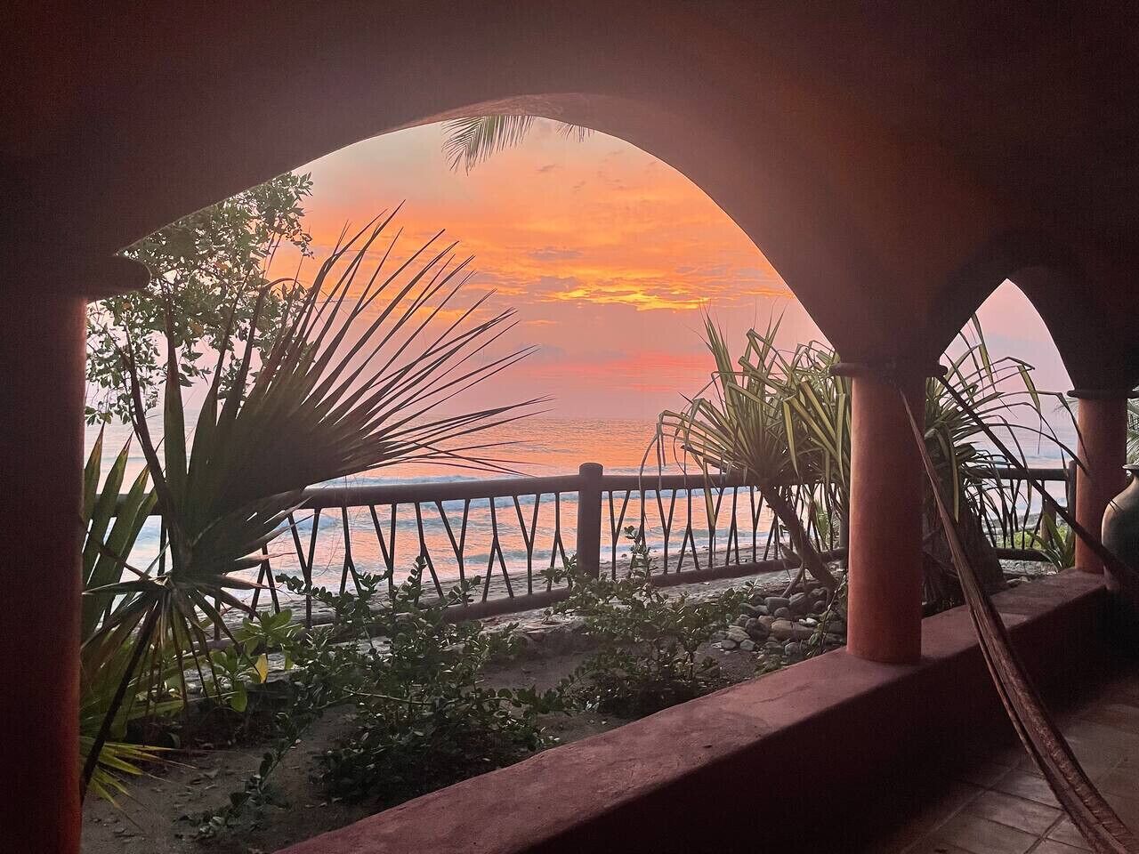A sunset view through an arched window overlooking a beach with a wooden railing and tropical plants.