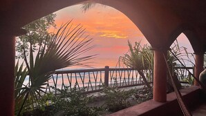 Vista al atardecer a través de un ventanal arqueado con vista a una playa, una barandilla de madera y plantas tropicales.