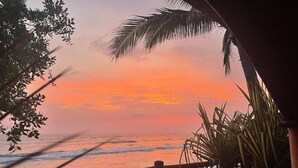 Vista del atardecer desde un balcón con baranda de madera, palmeras y olas del océano.