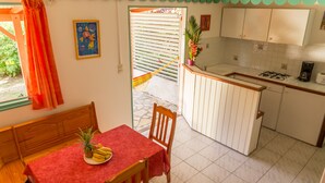 Una cocina con armarios blancos, un comedor con mantel rojo y vista a un jardín por la ventana.