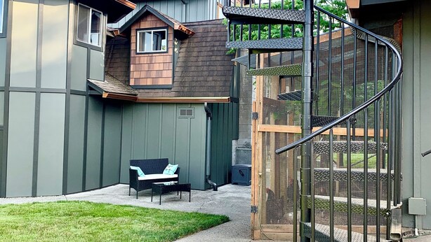 Un barrio residencial con una escalera en espiral que lleva a un balcón, un pequeño patio con mobiliario exterior, y una casa con techo inclinado.