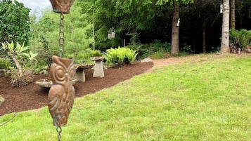 A garden with a hanging metal owl decoration, a stone birdbath, and a variety of greenery including ferns and trees.
