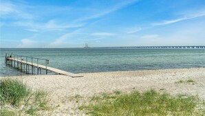 Een strand met een houten pier die het water in reikt, een heldere lucht en een lange brug in de verte.