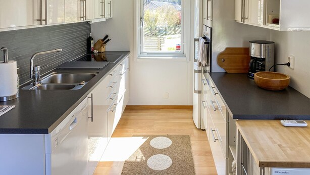 Una cocina moderna con encimera negra, pileta de acero inoxidable y muebles blancos. Se ve un ventanal con vistas a árboles y un edificio.