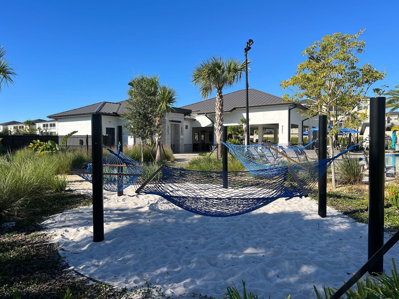 A hammock set up in a sandy area with a pool and a building in the background.