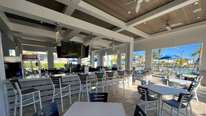 An outdoor dining area with white tables and chairs, a bar area with a television, and a view of a swimming pool and palm trees.