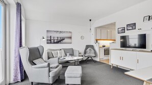 En moderne stue med sofasæt, et sofabord, et fjernsyn og et køkken der kan ses igennem en åben dør.