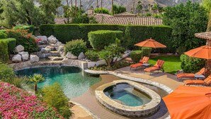 Una piscina con un jacuzzi, rodeada por un jardín ajardinado con rocas y plantas. Hay sillas de descanso y sombrillas.