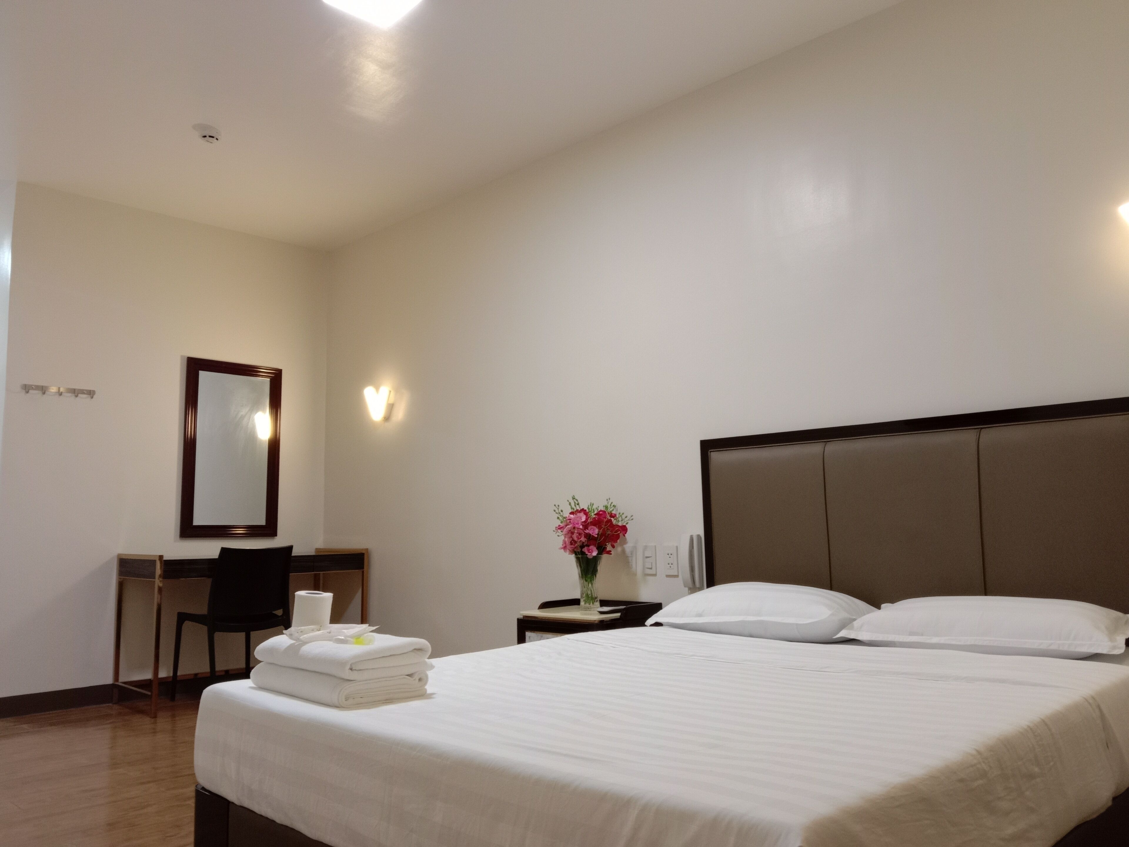 A hotel room with a large bed, a desk, a chair, a mirror, and a vase with flowers.