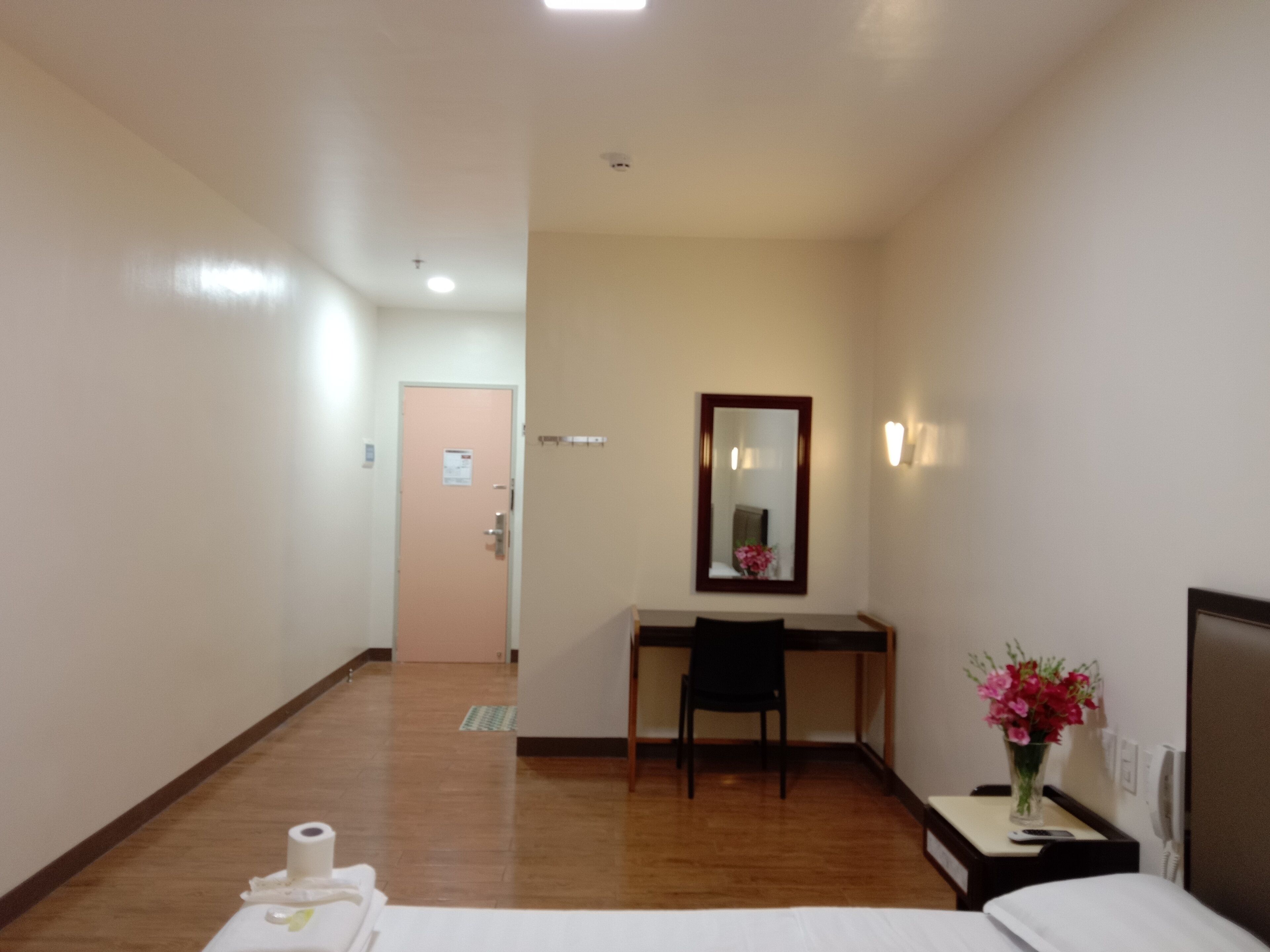 Habitación de hotel con cama, escritorio, silla, espejo, mesita de noche con un jarrón de flores y una puerta de baño al fondo.
