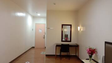 Habitación de hotel con cama, escritorio, silla, espejo, mesita de noche con un jarrón de flores y una puerta de baño al fondo.